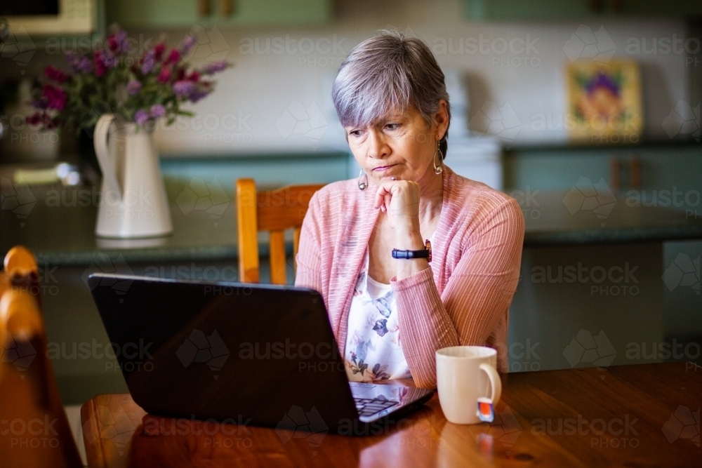 Senior woman with cup of tea working on laptop from home - Australian Stock Image