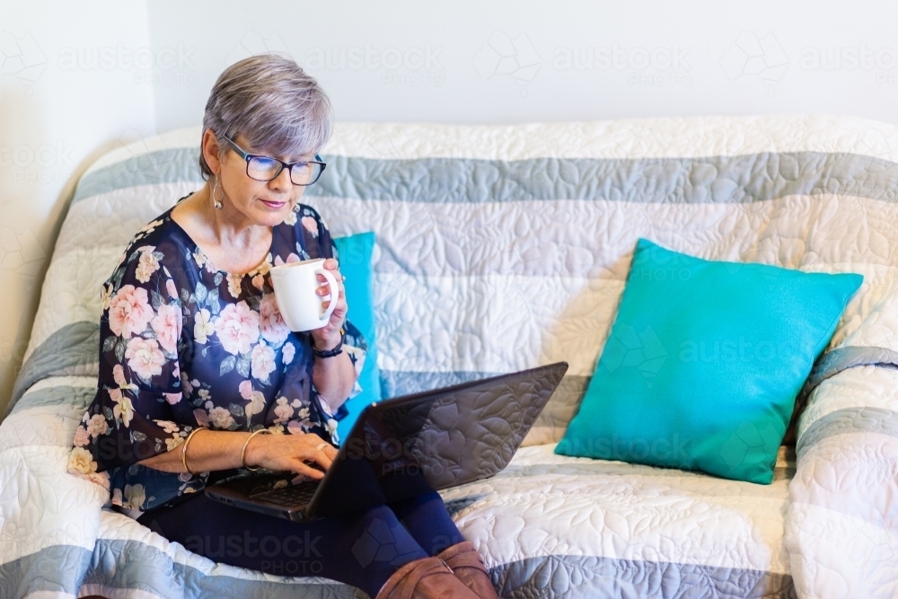 senior woman working on laptop on lounge with a cup of tea - Australian Stock Image