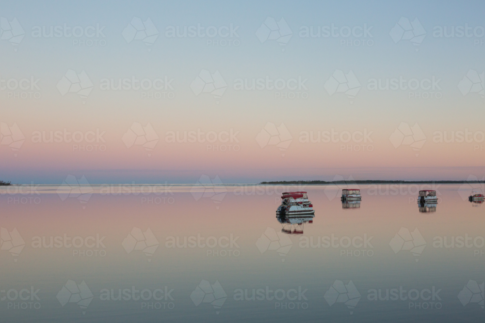 Serene twilight scene. Calm water mirrors pastel sky and small boats, peaceful, minimalist landscape - Australian Stock Image