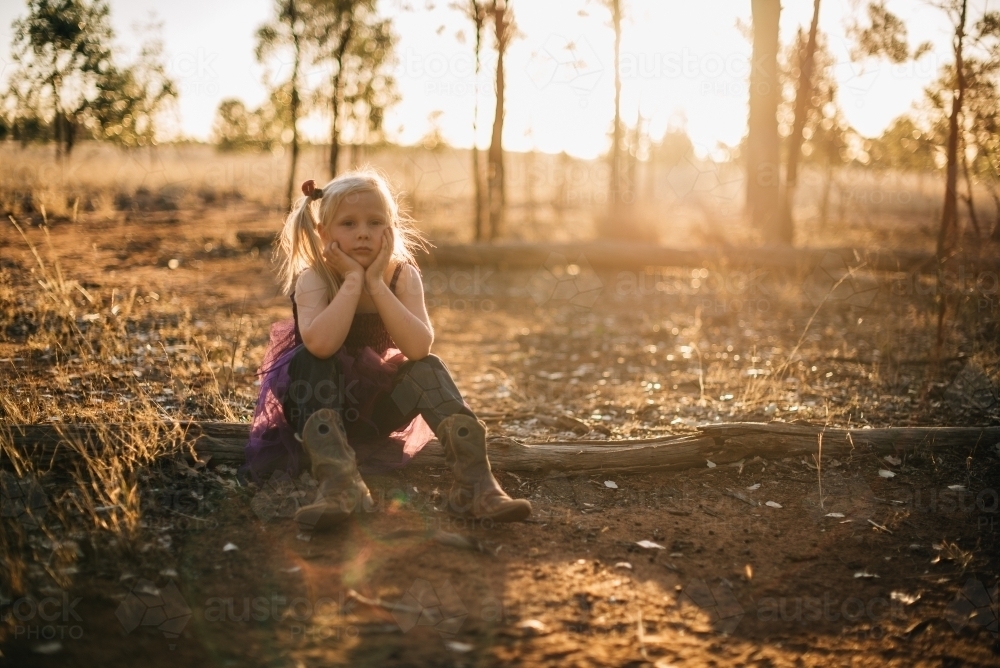 Serious young girl sitting outside with face leaning on hands as the sun sets - Australian Stock Image