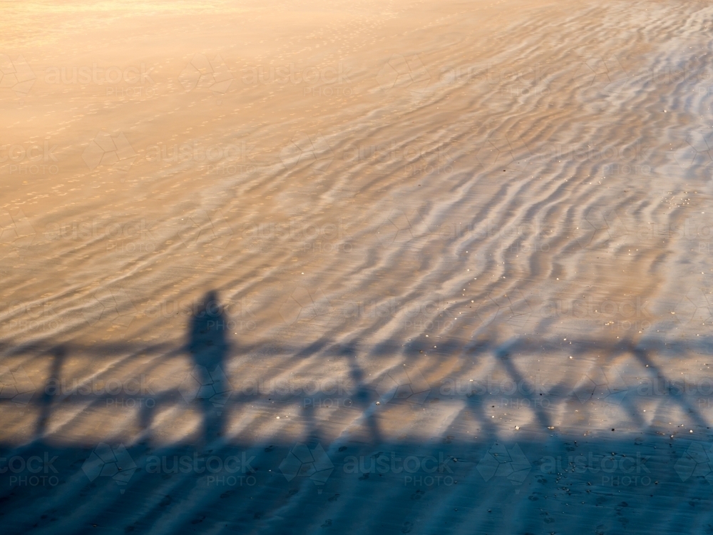 Shadow of a person, leaning on a rail, on a rippled beach sand pattern - Australian Stock Image
