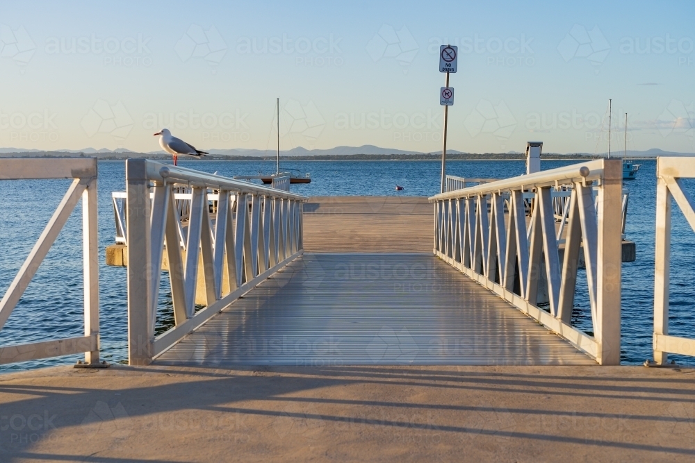 Shiny guard rails leading down onto a coastal jetty over a calm bay with blue sky overhead - Australian Stock Image