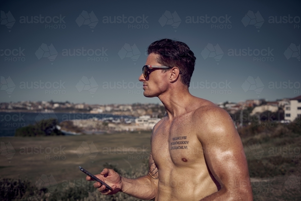 Shirtless male with phone beside ocean - Australian Stock Image