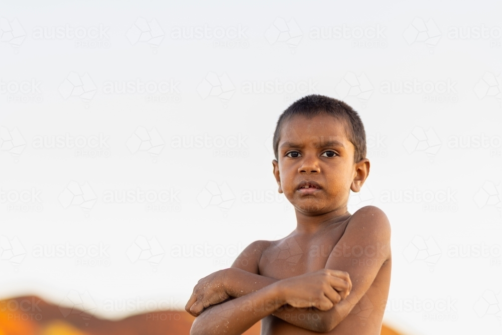 shirtless young aboriginal boy with sand on his skin against plain sky background - Australian Stock Image