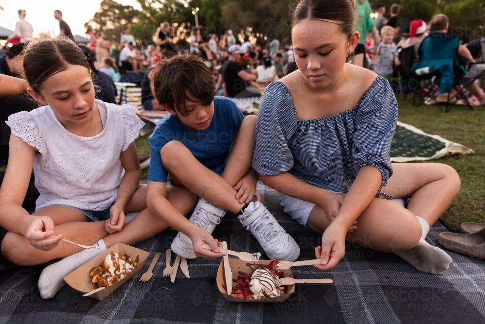 siblings sharing waffles on a picnic rug at the carols - Australian Stock Image
