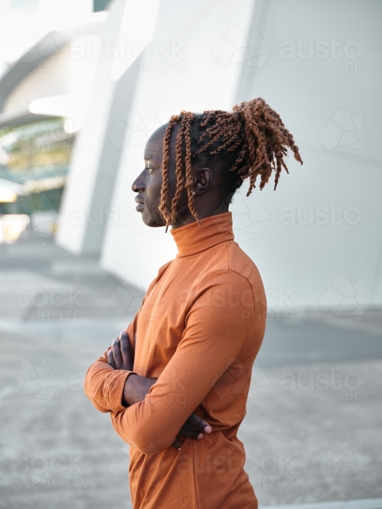 Side profile of South Sudanese man wearing orange turtleneck with arms folded - Australian Stock Image