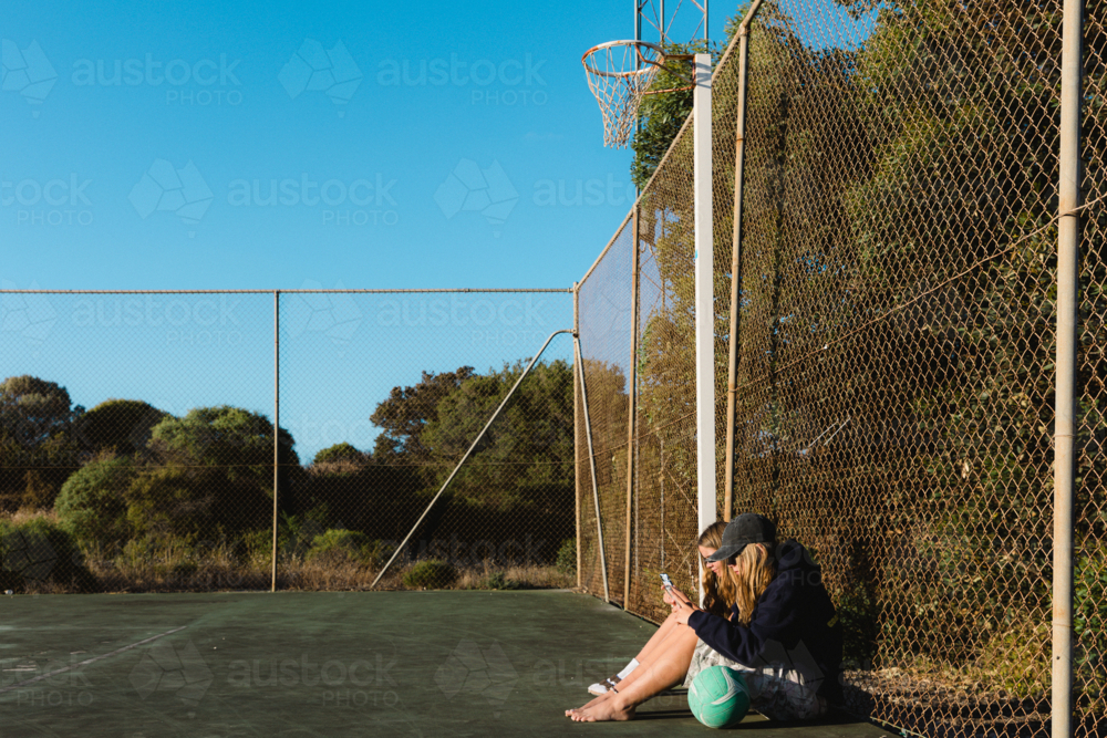 Side view of teenage girls on smart phones with a ball at outdoor netball court - Australian Stock Image