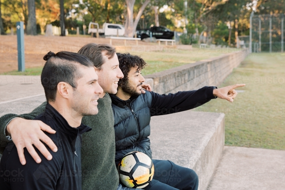 Side view photo of 3 smiling interracial man sitting on a field, one is pointing a finger - Australian Stock Image