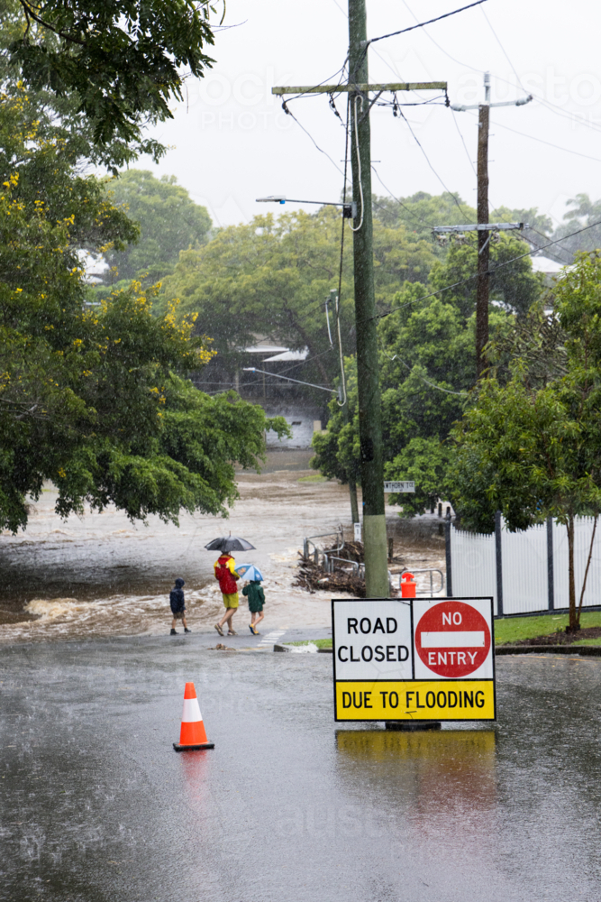 Signage across a flooded suburban road with pedestrians in background with umbrellas - Australian Stock Image