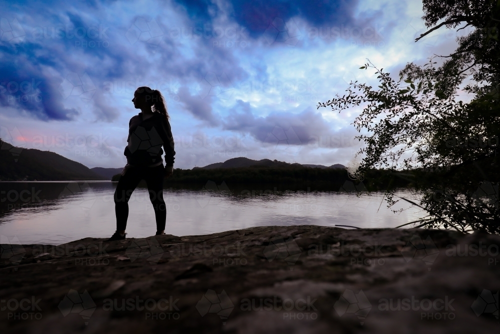 Silhouette image of woman standing on rock by river at twilight. - Australian Stock Image