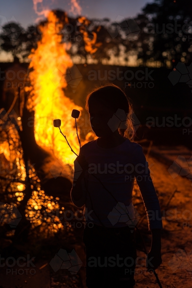 silhouette of a kid with marshmallows on a stick by a bonfire - Australian Stock Image