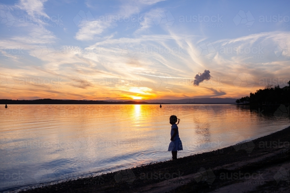 Silhouette of a little girl child standing at the edge of a lake at sundown - Australian Stock Image