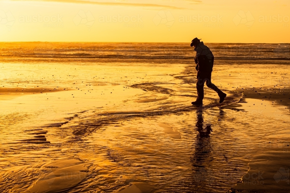 Silhouette of a man on a golden sunset lit beach with golden reflections - Australian Stock Image