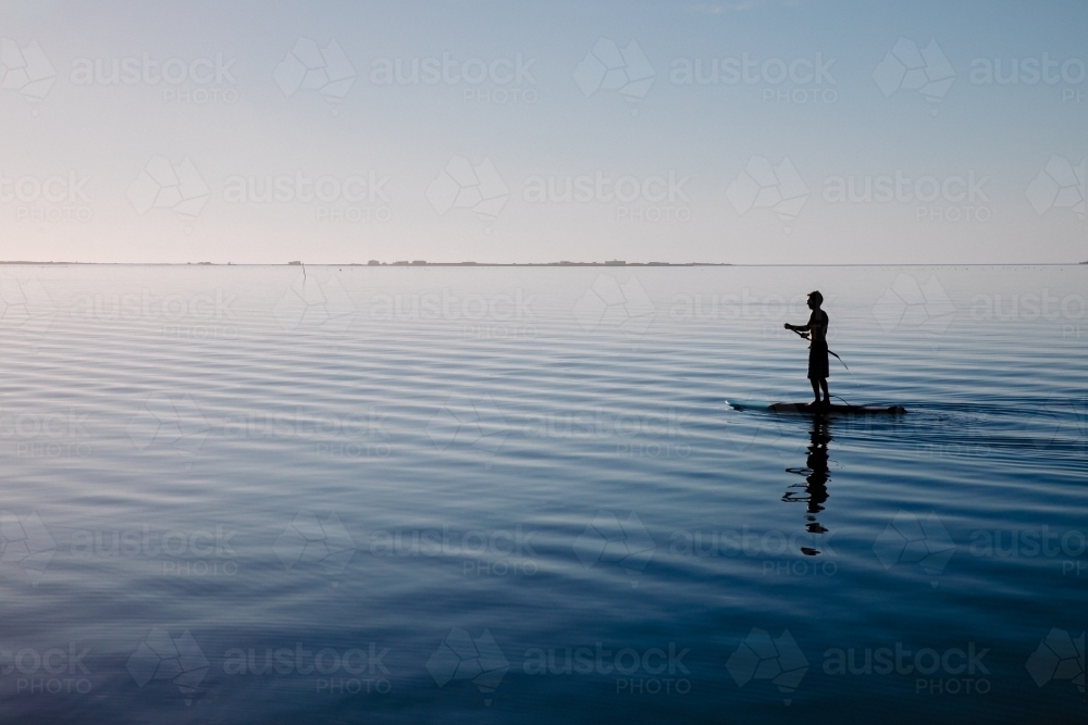 Silhouette of a man on a stand-up paddle board on very still water - Australian Stock Image