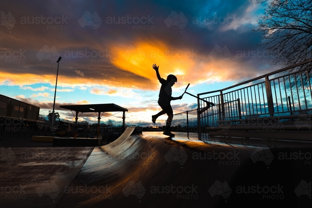 Silhouette of boy riding scooter on skate park ramp at sunset in Mansfield, Victoria - Australian Stock Image