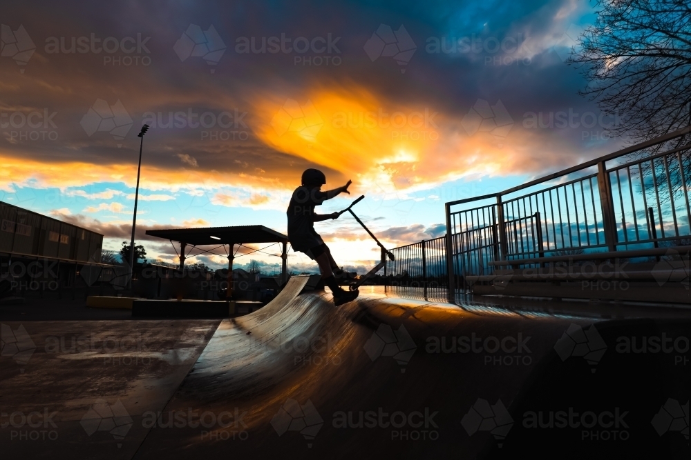 Silhouette of boy riding scooter on skate park ramp at sunset in Mansfield, Victoria - Australian Stock Image