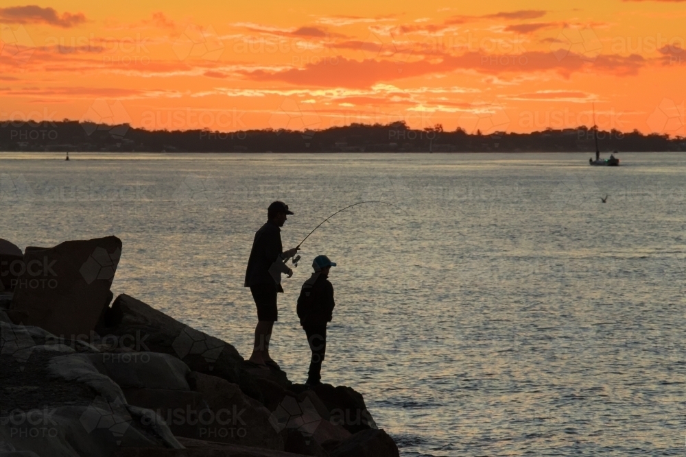 Silhouette of father and son fishing off breakwall at sunset - Australian Stock Image
