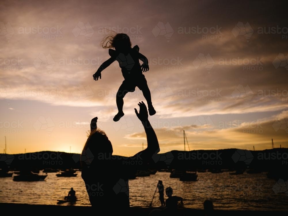 Silhouette of father throwing daughter into the air at sunset - Australian Stock Image