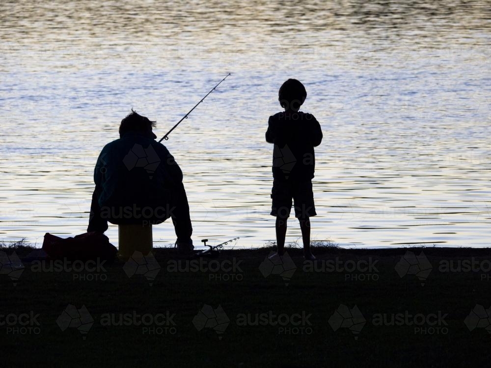 Silhouette of fisherman and boy at water's edge - Australian Stock Image