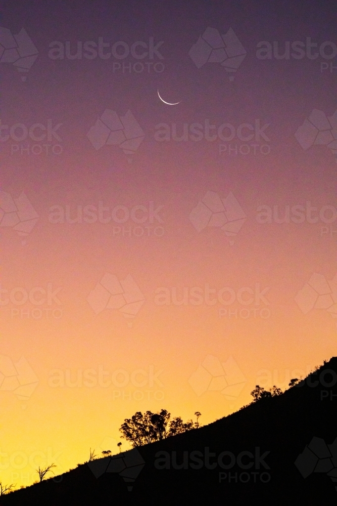 silhouette of hillside against dusk sky with crescent moon - Australian Stock Image