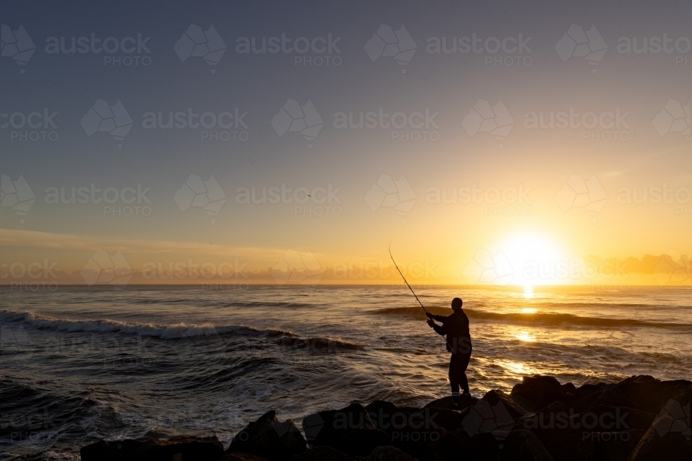 Silhouette of man fishing off rocks at sunrise - Australian Stock Image