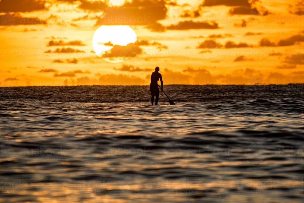 Silhouette of paddler with the large sun rising behind him on the ocean water - Australian Stock Image