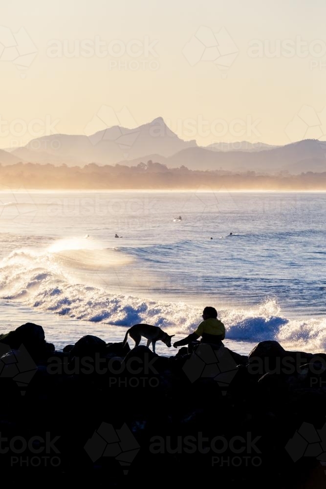 Silhouette of person and dog on rocks watching surfers at Byron Bay - Australian Stock Image