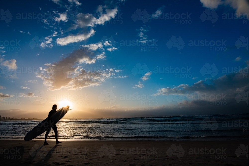 Silhouette of surfer carrying his surfboard to the water along the shore - Australian Stock Image