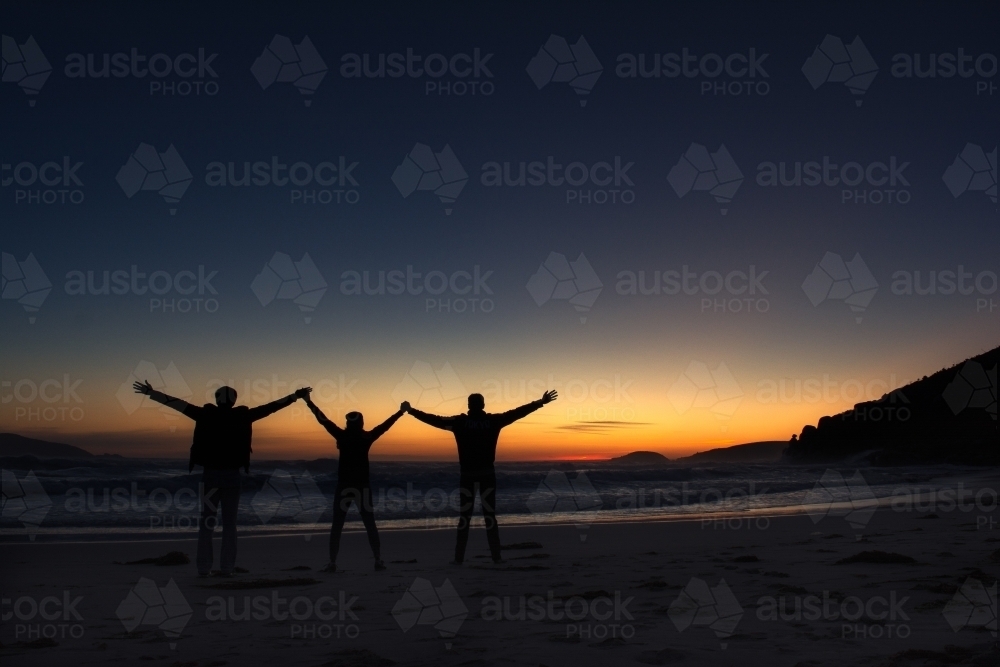 Silhouette of three friends holding hands raising against a sunset background - Australian Stock Image