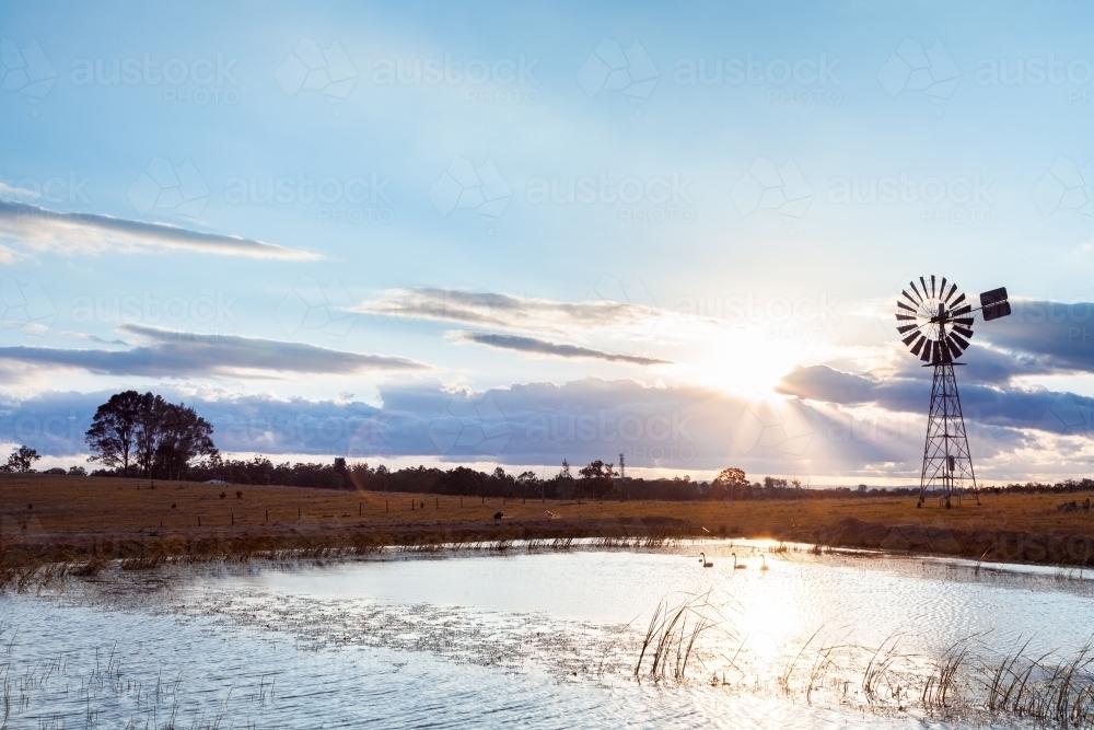 Silhouette of windmill and sunlight and farm dam at sunset - Australian Stock Image
