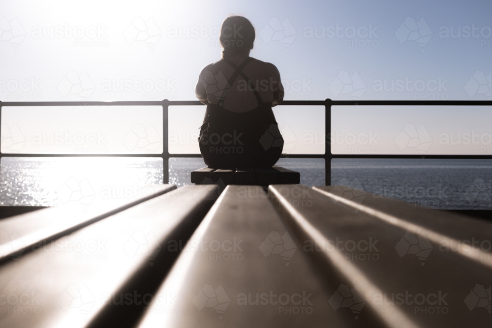Silhouette of woman sitting on jetty bench looking out to sea - Australian Stock Image