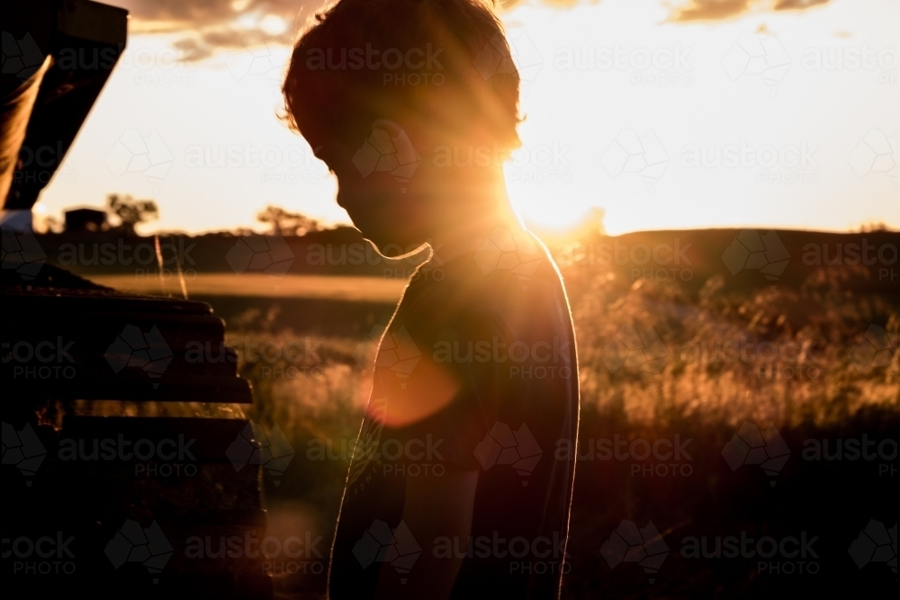 Silhouette of young boy & machinery, rural farming property in background as sun setting - Australian Stock Image