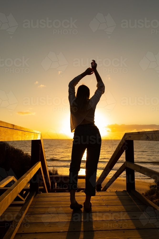 silhouette of young woman with arms in the air celebrate life against sunset at the beach - Australian Stock Image