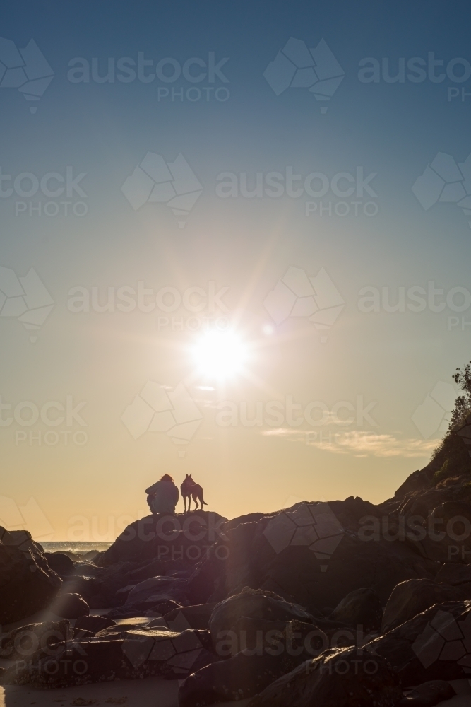 Silhouette person and dog sitting on rocks at sunset - Australian Stock Image