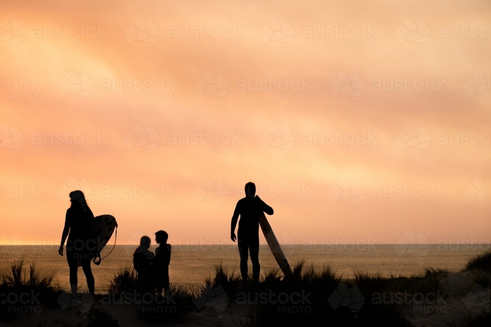 silhouette portrait of surfing family of four overlooking ocean on coastal dune holding surfboards - Australian Stock Image