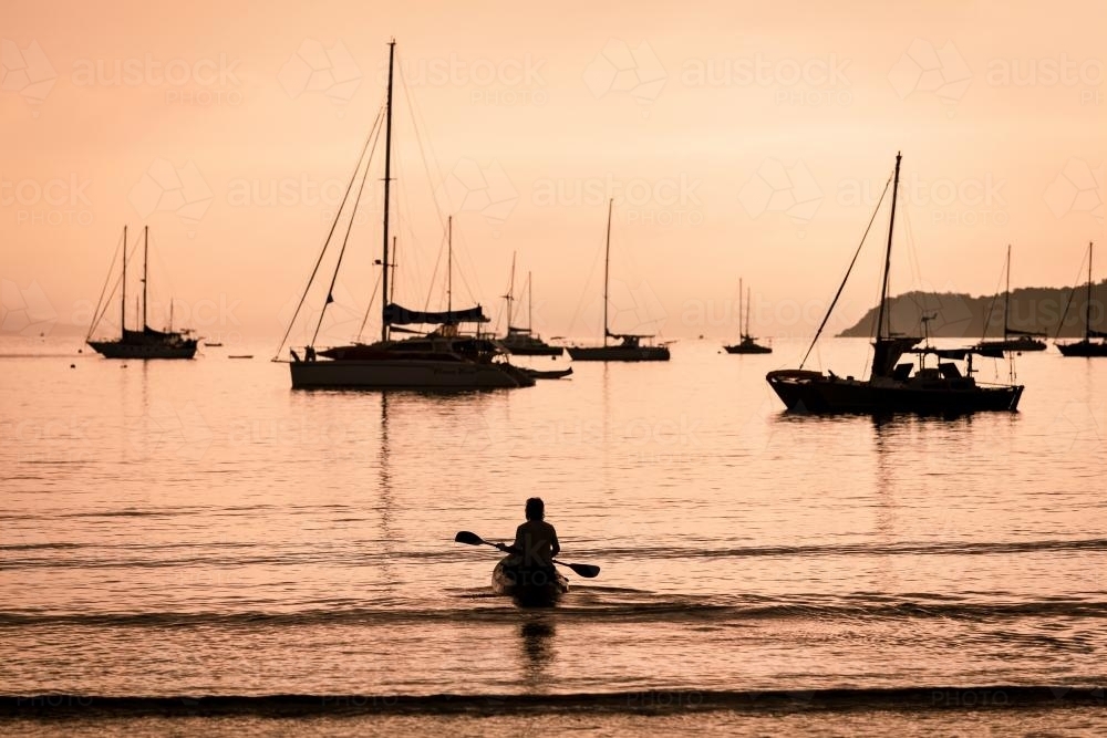 Silhouetted boats and person kayaking on a calm morning off Airlie Beach - Australian Stock Image