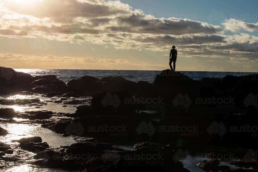 Silhouetted female standing on rocky headland early in the morning - Australian Stock Image