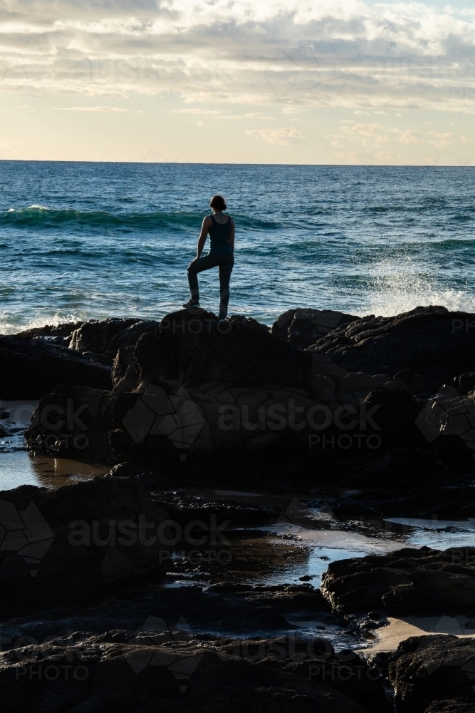 Silhouetted female standing on rocky headland early in the morning - Australian Stock Image