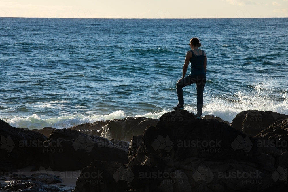 Silhouetted female standing on rocky headland early in the morning - Australian Stock Image