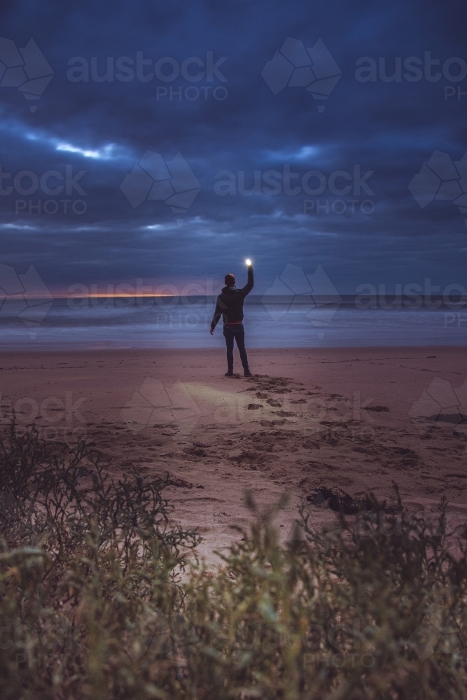 Silhouetted Figure Holding Up a Light on a Dark Beach - Australian Stock Image