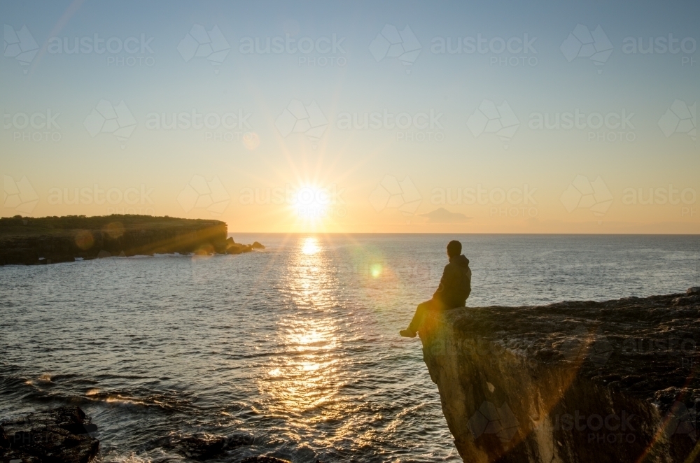 Silhouetted man on a cliff watching the sunrise by the sea - Australian Stock Image