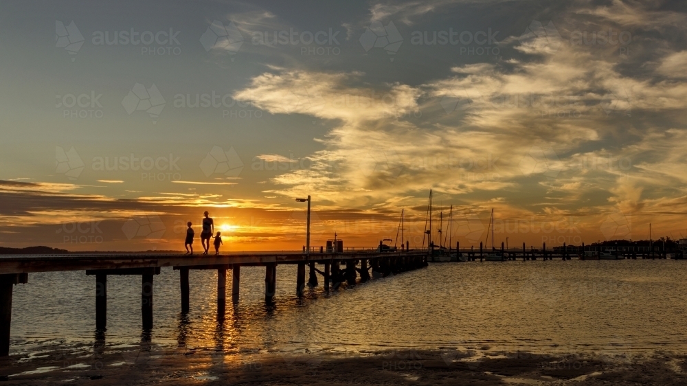 Silhouetted mother & her 2 daughters walking along wooden jetty at sunset - Australian Stock Image