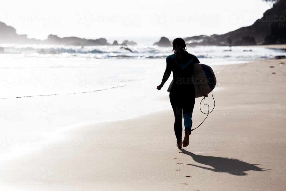 Silhouetted surfer running on beach with surfboard in bright summer light - Australian Stock Image