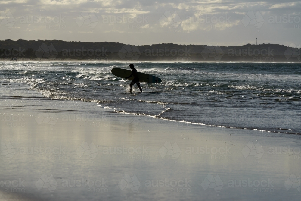 Silhouetted surfer walking out of the water carrying his board with glistening water - Australian Stock Image