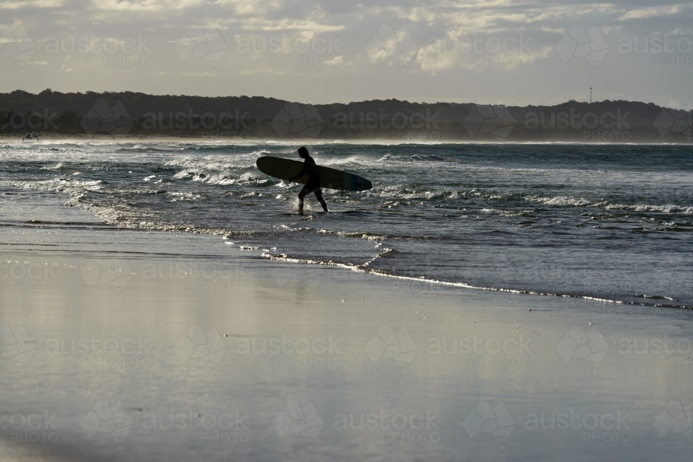 Silhouetted surfer walking out of the water carrying his board with glistening water - Australian Stock Image