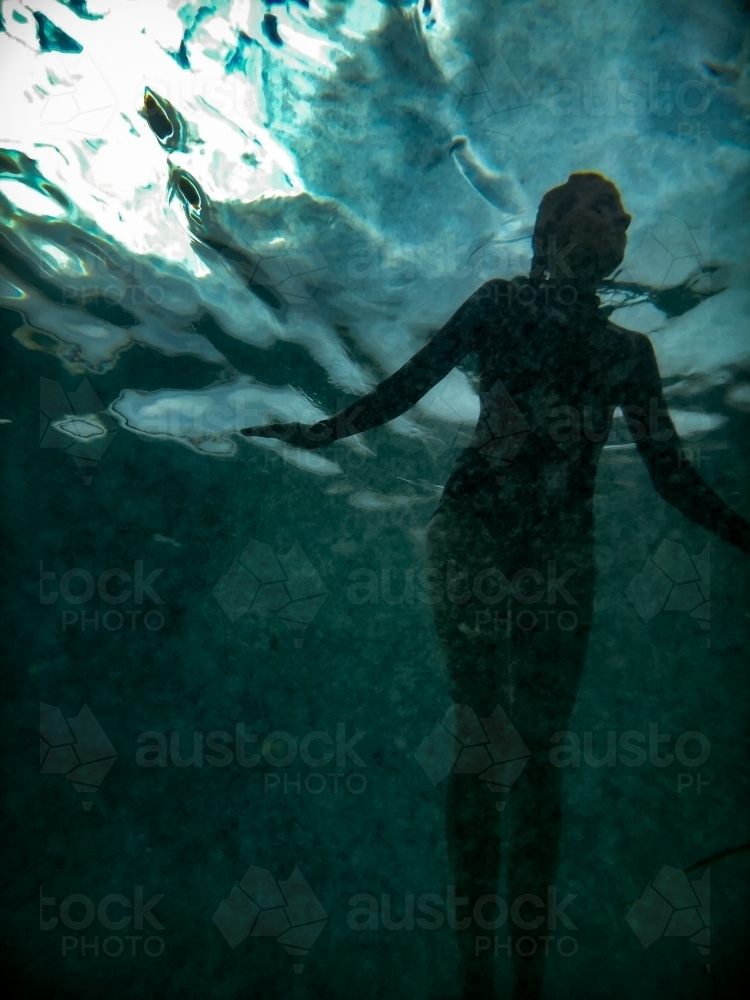 Silhouetted swimming female figure looking upwards underwater towards ocean surface - Australian Stock Image