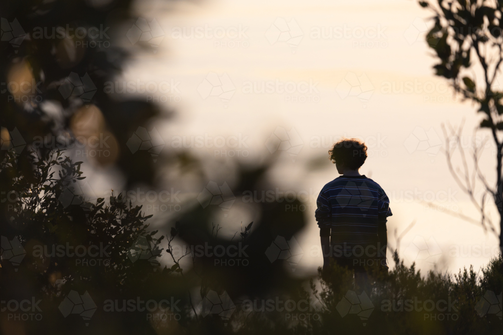 Silhouetted teenage woman looking at sunset view of calm sea while bush walking in nature - Australian Stock Image