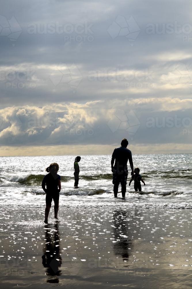 Silhouettes of a family paddling in the shallows on a sunlit beach - Australian Stock Image