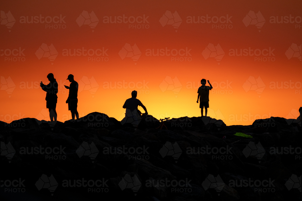Silhouettes of people on rocks against a vivid orange and yellow sunset sky. - Australian Stock Image