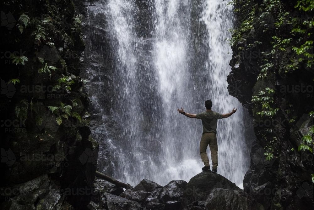Single man with arms lifted in front of waterfall - Australian Stock Image